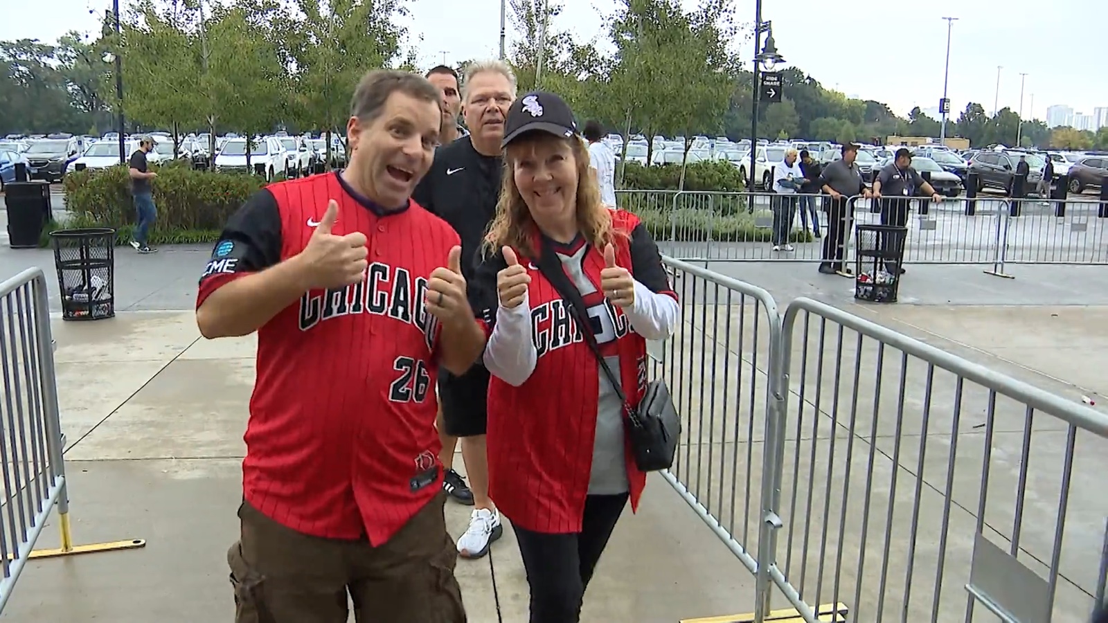 A man and a woman in red Chicago White Sox jerseys give thumbs up outside the ballpark, the woman in a black Sox cap, a car park and trees behind them