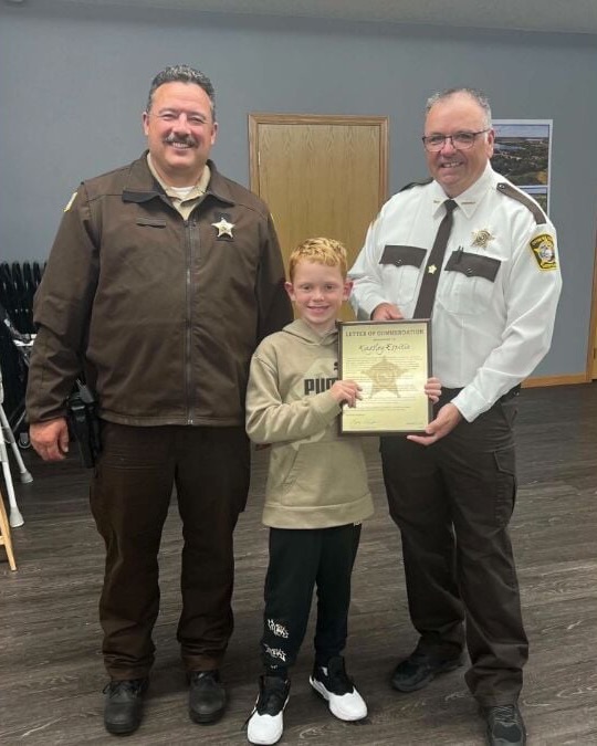 Kingsley Espitia, a small boy with red hair in a beige hoodie and white trainers, holding a framed letter of commendation between two Otter Tail County sheriff's officers in a council room