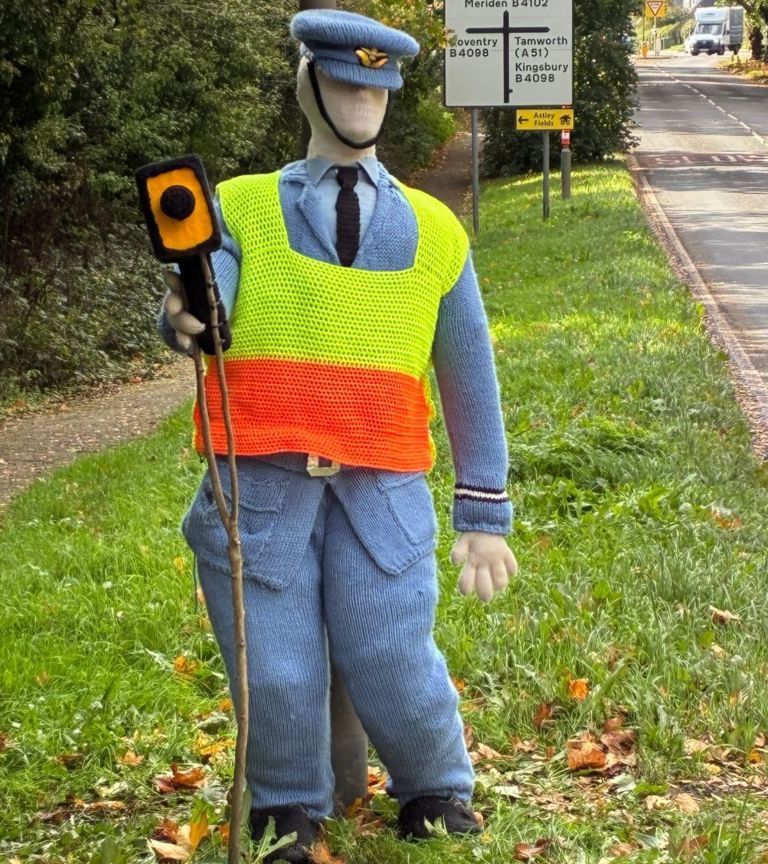 A life-sized knitted figure in a blue RAF uniform, cap and orange and yellow hi-vis vest stands on a grass verge, holding a knitted speed gun towards the road.