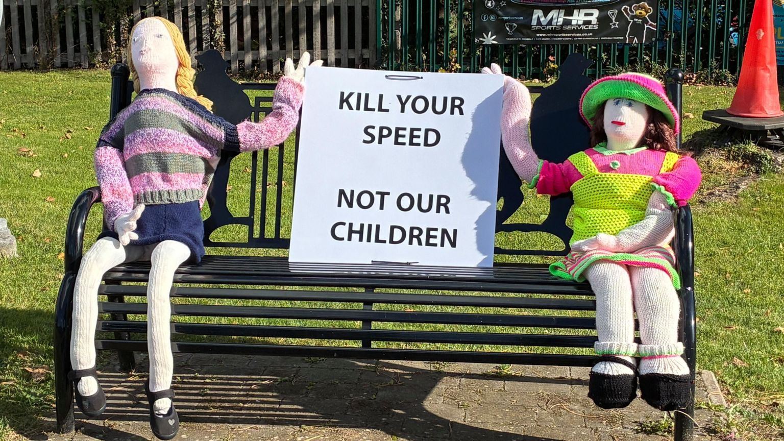 Two knitted children sit on a bench holding a sign that reads Kill your speed, not our children.