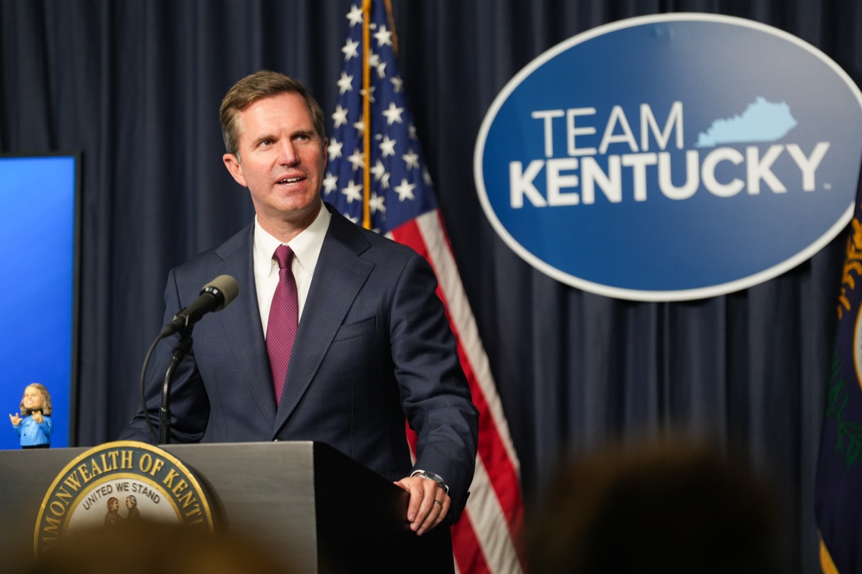 Governor Andy Beshear speaking at a podium bearing the Commonwealth of Kentucky seal, with a Team Kentucky sign behind him
