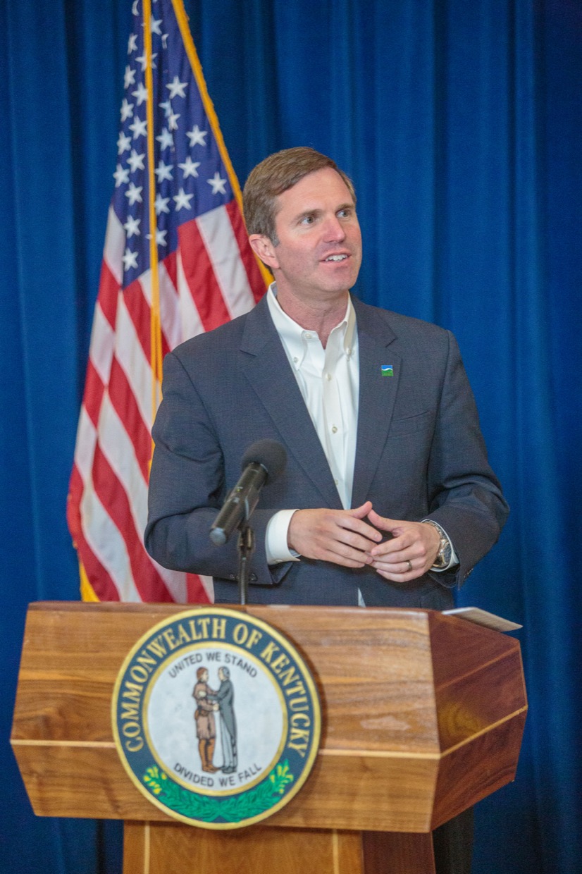 Governor Andy Beshear speaking at a podium bearing the Commonwealth of Kentucky seal, an American flag behind him