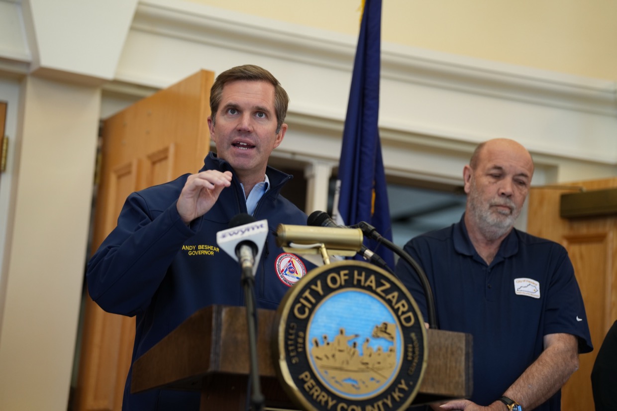 Governor Beshear in a windbreaker speaking at a City of Hazard podium, a local official standing beside him