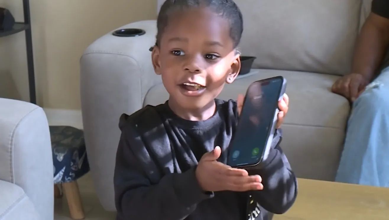 A small boy in a black top sits on a sofa holding up a phone and talking.