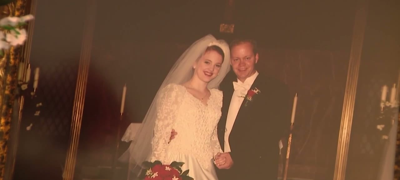 A framed wedding photograph: a bride in a white lace gown and veil holding red roses, beside a groom in a black tuxedo and white bow tie, both smiling, candles behind them.