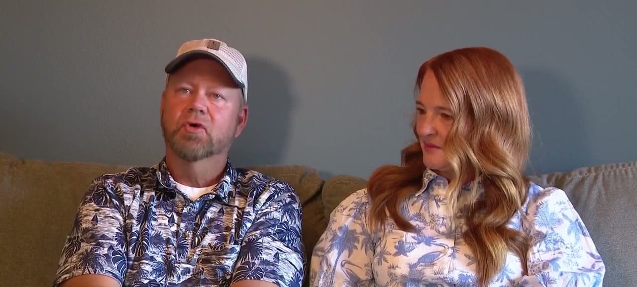 A man in a cap and a floral shirt sits on a sofa next to a woman with long red hair in a floral blouse, both talking to the camera.