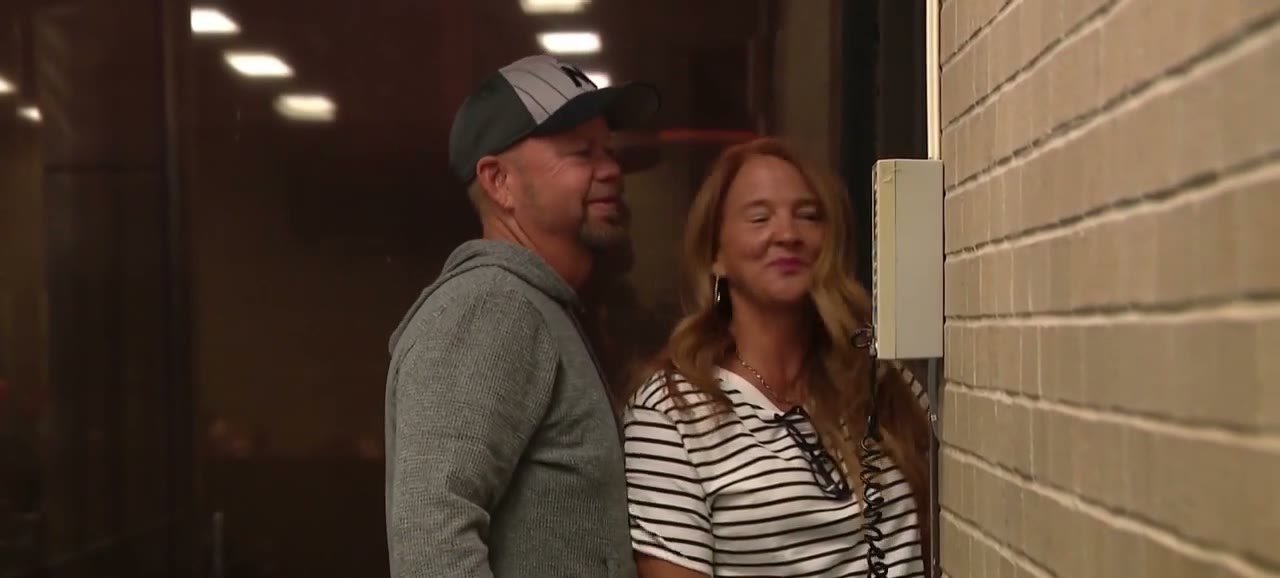 A man in a grey hoodie and dark cap walks beside a smiling woman in a striped top through an airport corridor, lit by overhead lights.