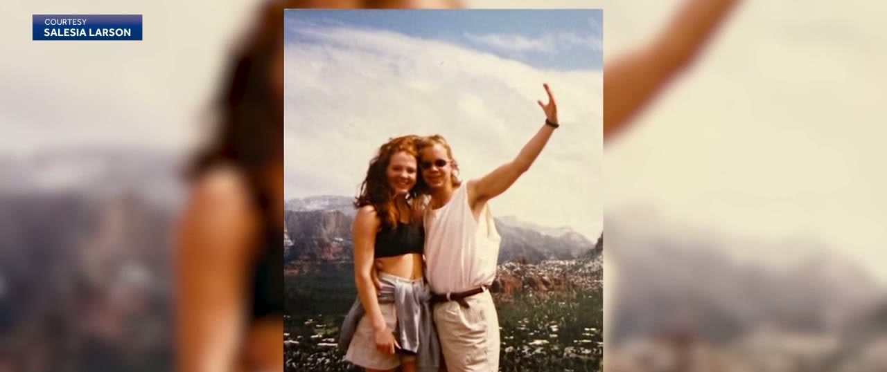 An old snapshot of a young couple in the mountains: a woman with long red hair in a dark top and a man in a white T-shirt with his arm raised, both smiling.