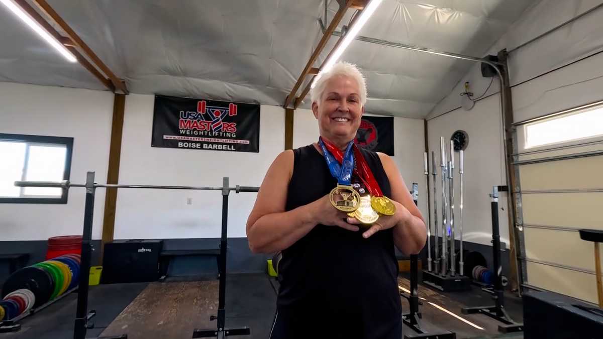 A smiling woman with short white hair holds three gold medals in a weightlifting gym, with bars and plates behind her.