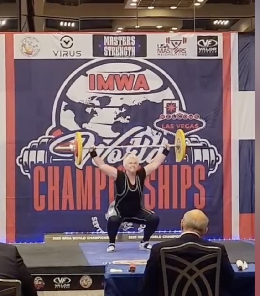A white-haired woman holds a loaded barbell overhead in a deep squat on a competition stage under a World Championships banner.