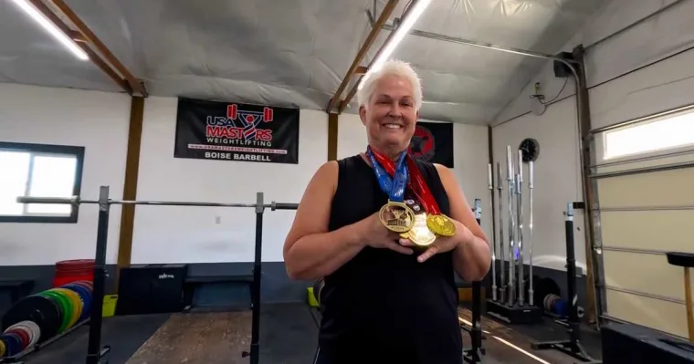 A smiling woman with short white hair holds three gold medals in a weightlifting gym, with bars and plates behind her.