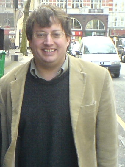 David Mitchell, smiling, in glasses, a beige corduroy jacket and a dark jumper, on a London street