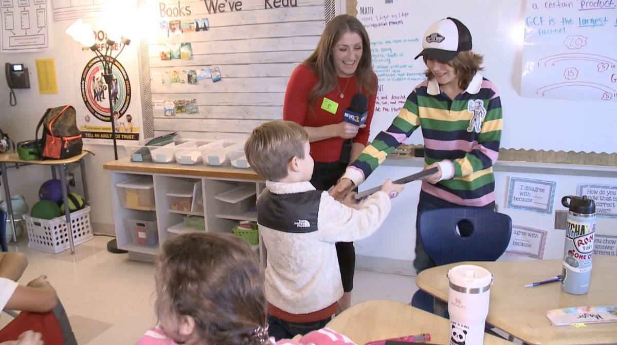 A television reporter holding a microphone in a classroom as Bryce Rader hands Major Metzger an award
