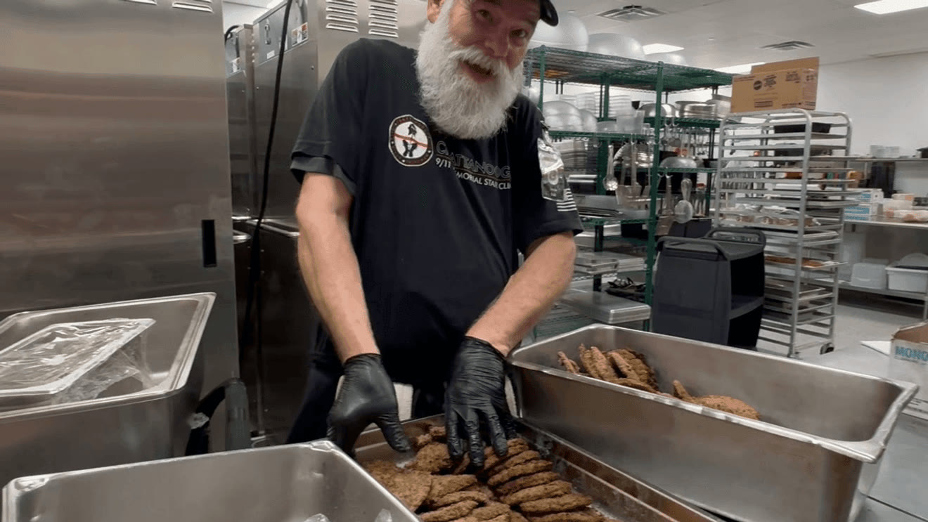 A bearded man in a black T-shirt, cap and black gloves leans over steel trays of cooked burger patties in a stainless-steel stadium kitchen, smiling at the camera