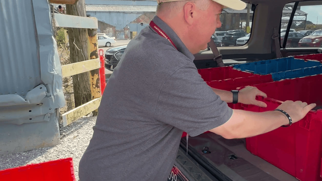 A man in a grey polo shirt and lanyard pushes red and blue plastic crates into the open back of a car in a gravel car park