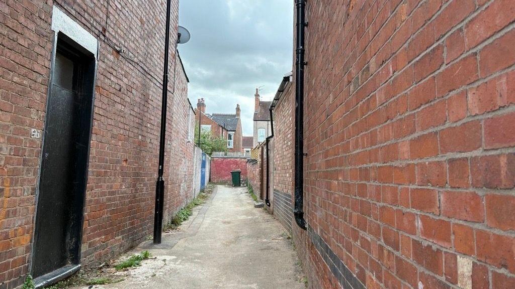The same alleyway on Friday, swept empty, red brick walls on both sides, a drainpipe, and a green wheelie bin at the far end under a grey sky