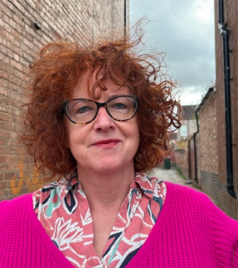 Mary Courtney standing in a narrow brick alleyway in Coventry, with curly red hair and dark-framed glasses, wearing a bright pink cardigan over a patterned shirt