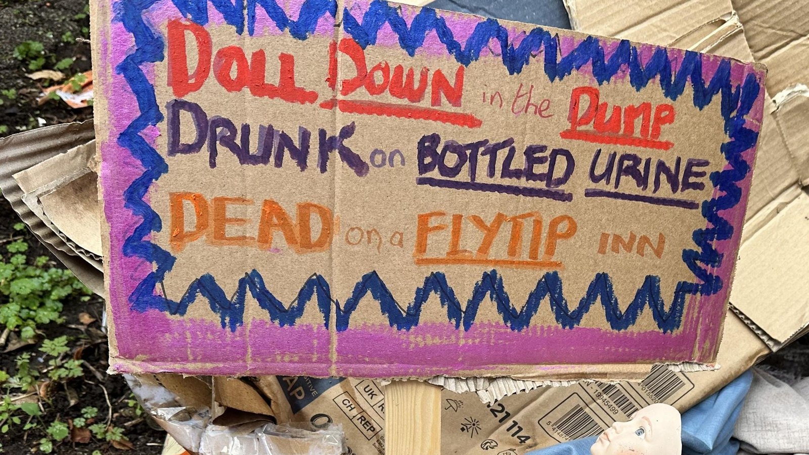 A piece of brown cardboard with a hand-lettered poem in red, purple and orange marker inside a blue zigzag border on magenta, reading Doll down in the dump, drunk on bottled urine, dead on a flytip inn