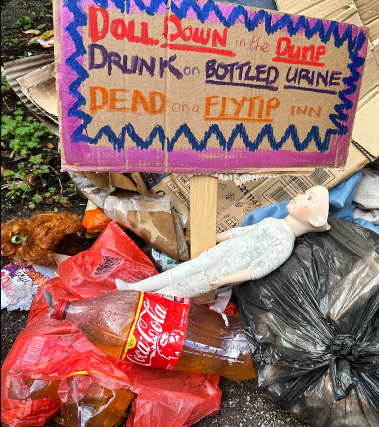 The fly-tip in the alley: a doll lying face up among black bin bags and torn cardboard, a Coca-Cola bottle filled with brown liquid in the foreground, and the cardboard protest sign standing above it all