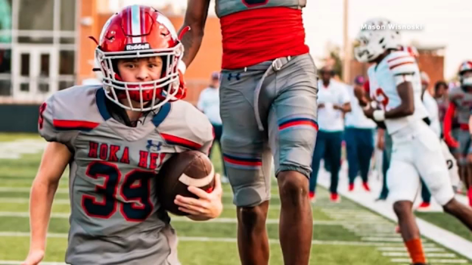 Mason Wisnoski runs with the ball tucked under his right arm, in a grey Manvel jersey number 39 with HOKA HEY across the chest and a red helmet, a teammate beside him with an arm raised