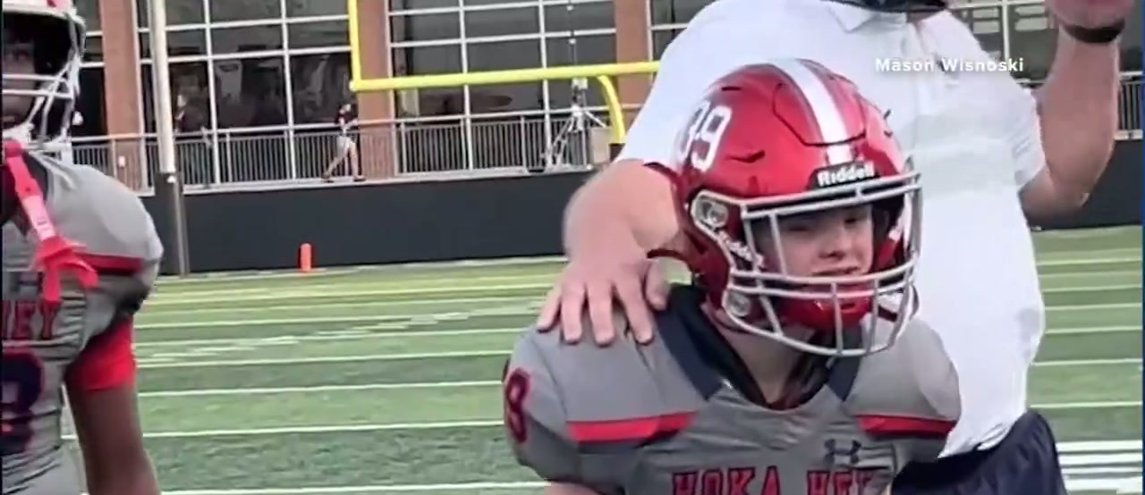 A teammate with his arm around Mason Wisnoski, who is still in his red helmet and grey HOKA HEY jersey, looking out across the field