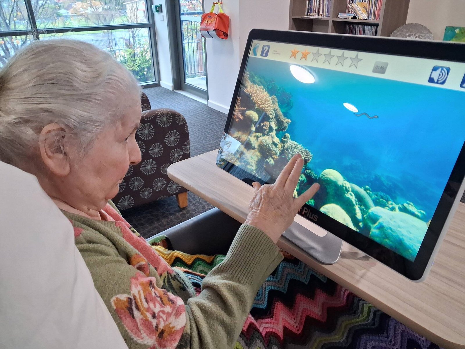 Noeleen, an older woman with white hair and a crocheted rug over her lap, reaches out to touch a large touchscreen showing a coral reef, in a lounge at Mercy Place Warrnambool