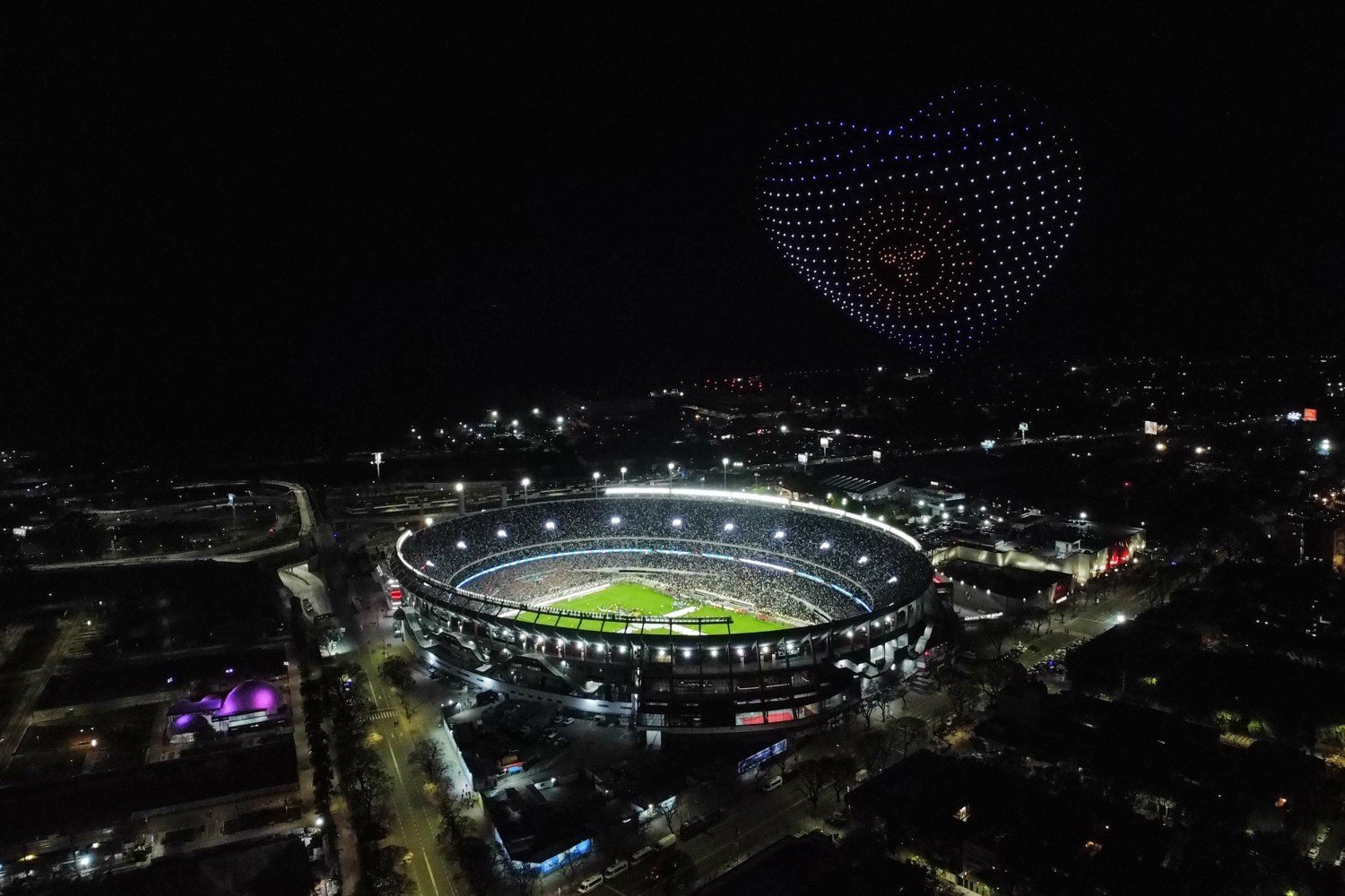 An aerial view at night of the floodlit Monumental stadium in Buenos Aires, with a heart made of drones in the sky above it.