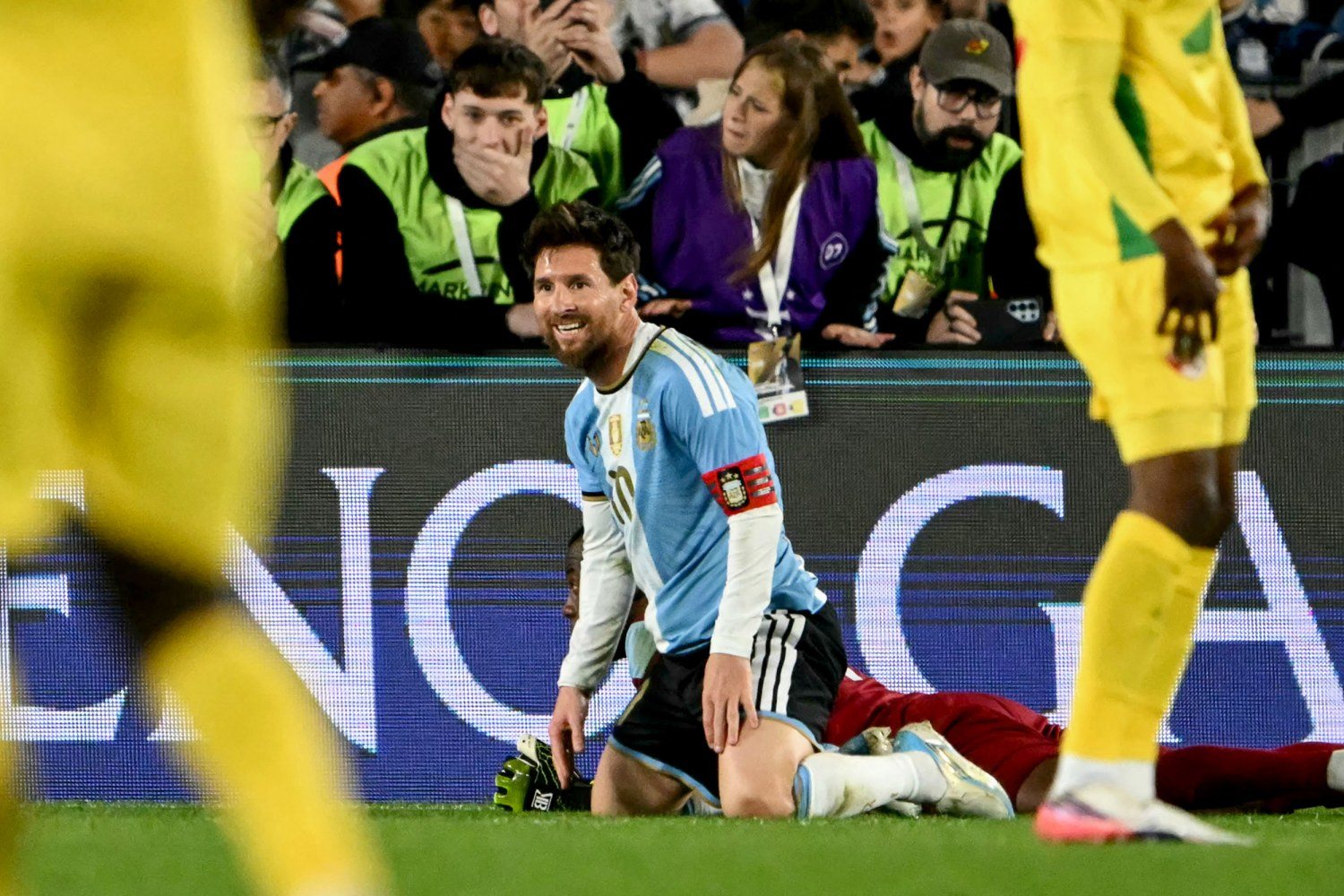 Messi kneeling on the grass in his number 10 shirt, looking up and smiling, with Benin players in yellow standing nearby.