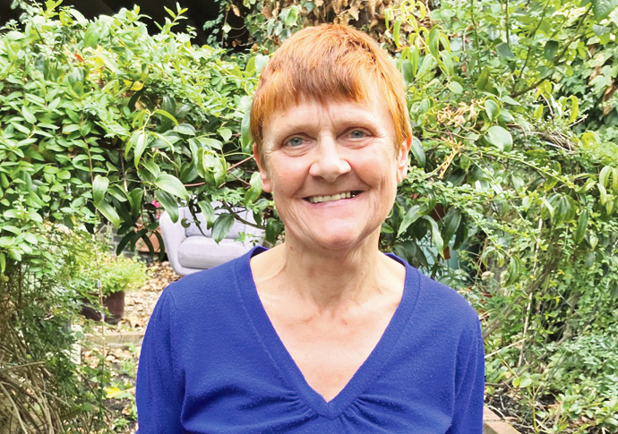 A smiling woman with short red hair in a blue V-neck top stands in a leafy garden