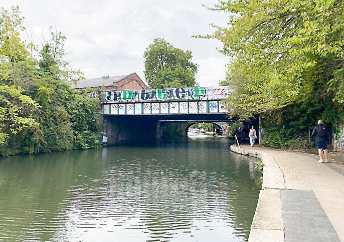 The Regent's Canal near Camden Lock on a grey day, still green water between overhanging trees, a graffitied iron bridge crossing ahead and a walker on the towpath to the right