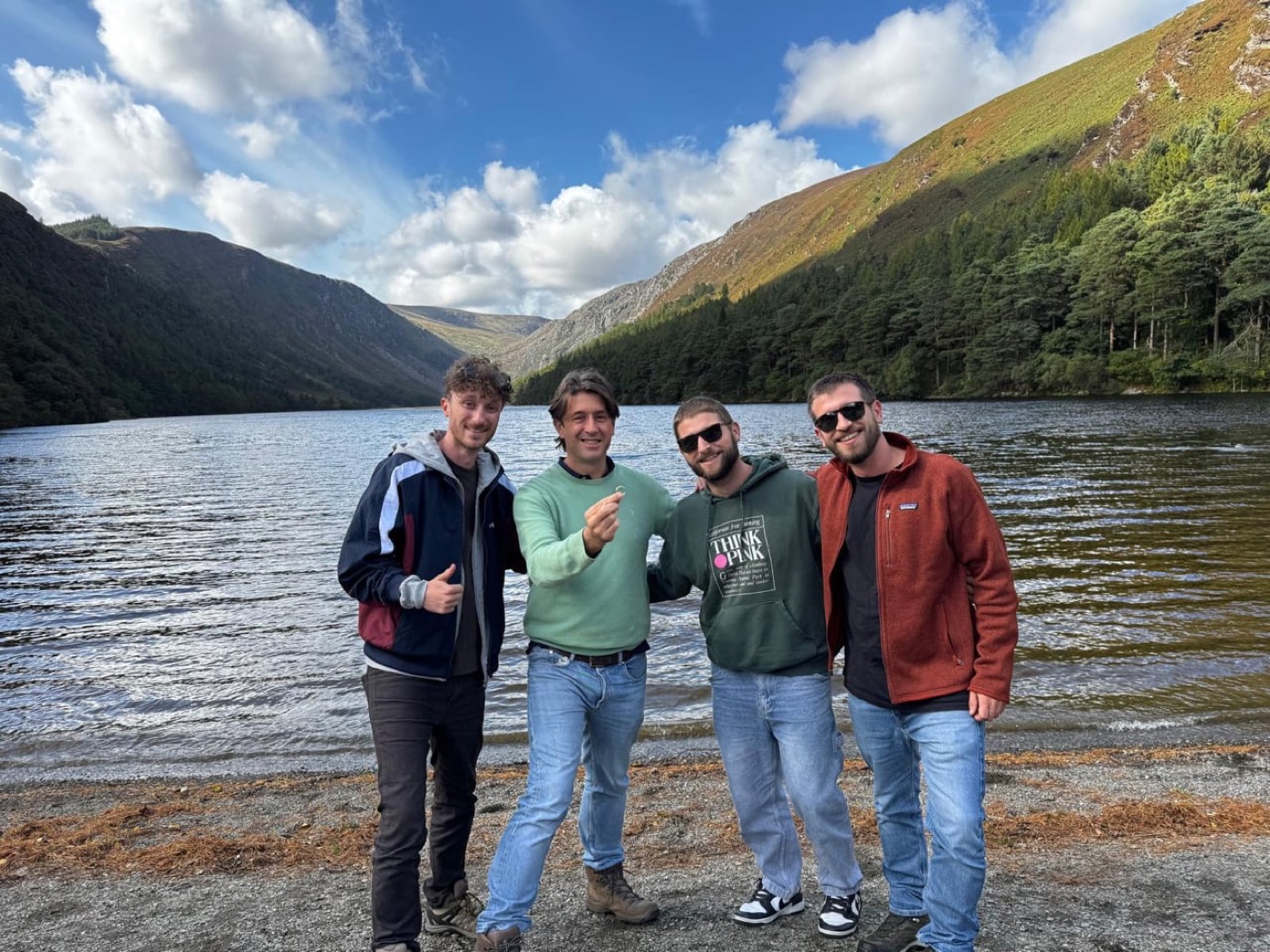 Four smiling men stand on a stony lake shore below green mountains. The second from left holds up a small gold ring.