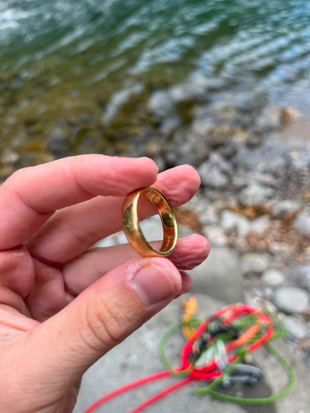A hand holds a gold wedding ring above a rocky stream bed, with red and green ropes coiled on the rocks below.