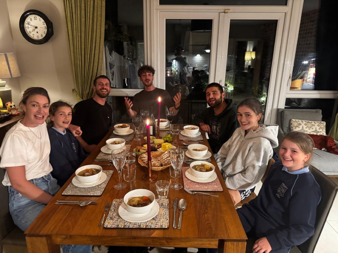 A family and three young men sit around a candlelit dinner table with bowls of stew, smiling at the camera.