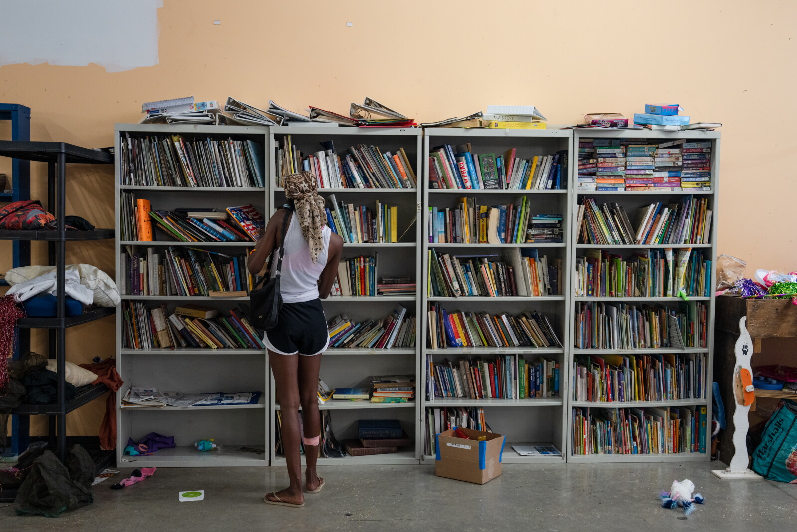 A woman with a headscarf stands looking at four tall shelving units packed with books, with more paperwork stacked on top and clothes on a rack to the left