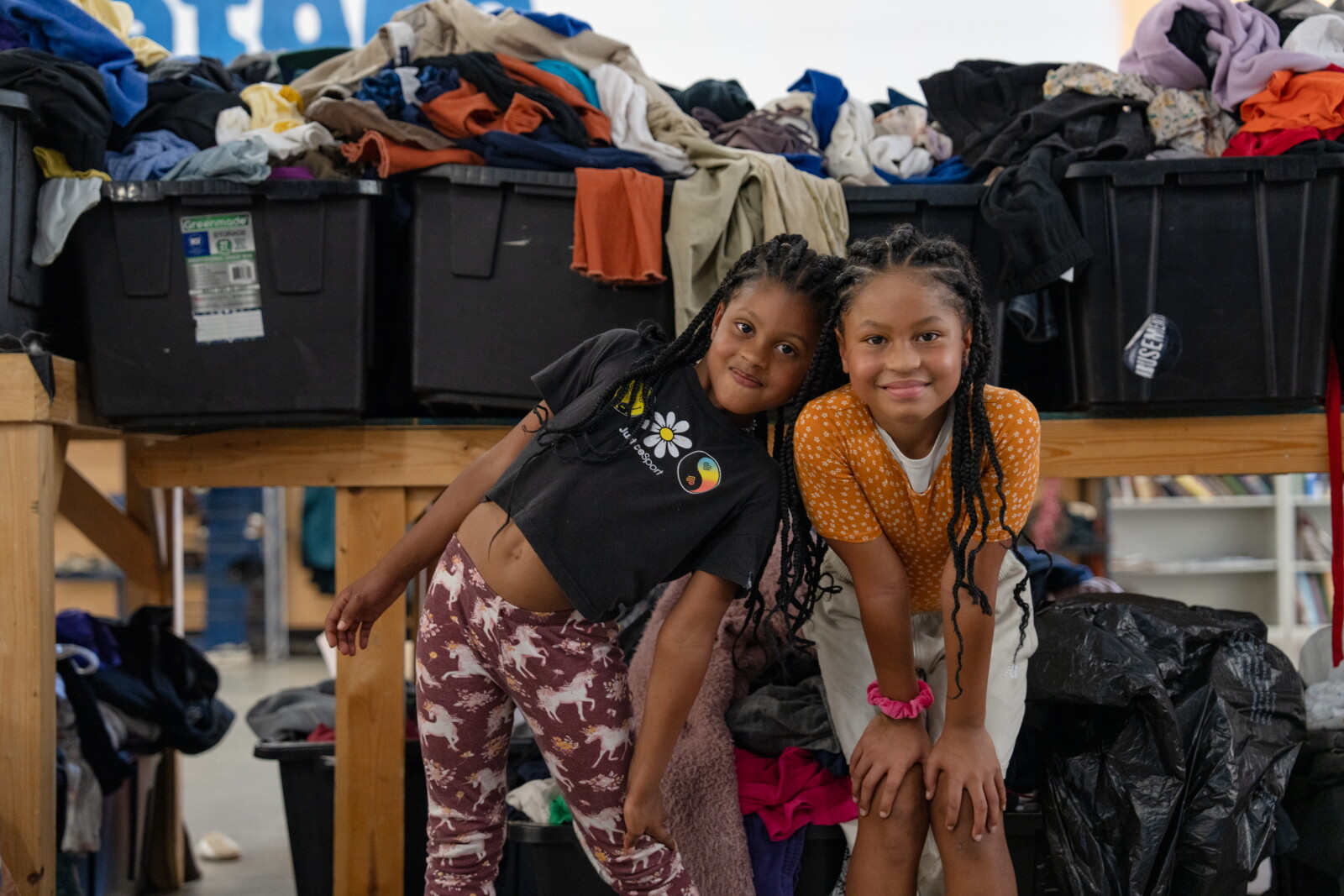 Two girls with braided hair lean together smiling in front of big plastic bins heaped with donated clothes