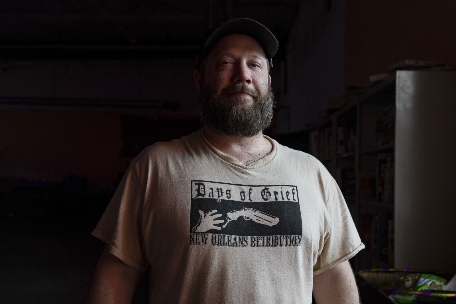 A bearded man in a flat cap and a pale T-shirt standing in the dim interior of the store, shelves behind him