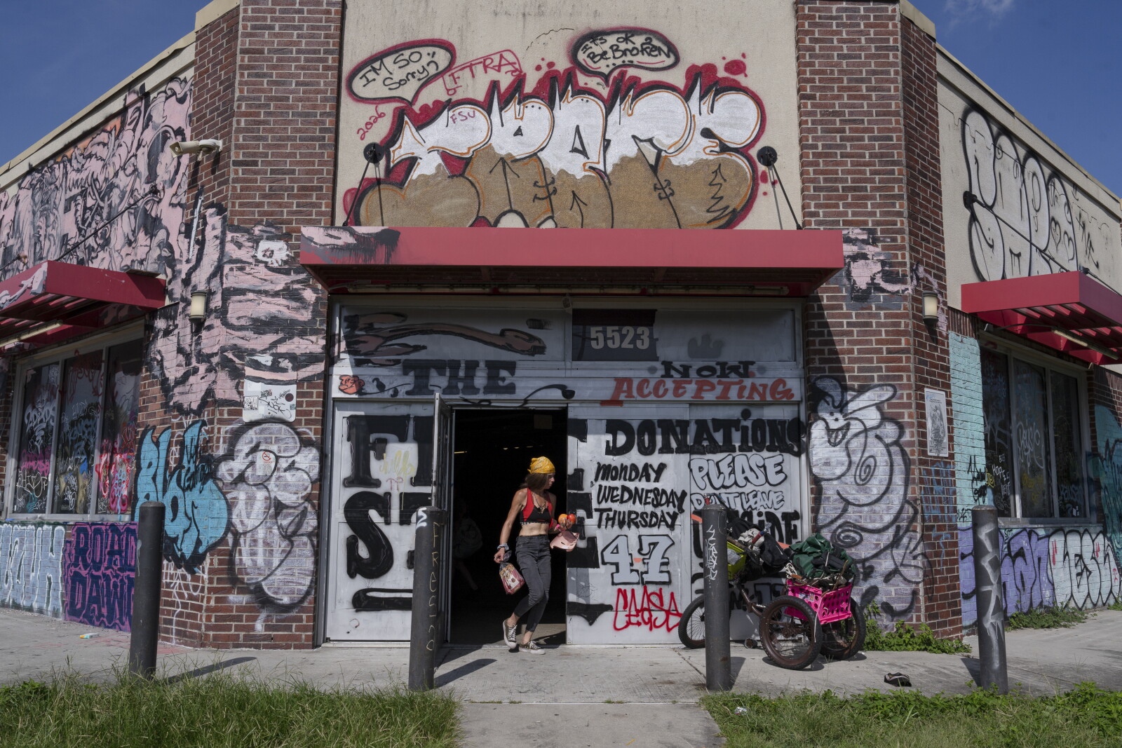 A graffiti-covered corner shop with red awnings, the words THE FREE STORE and NOW ACCEPTING DONATIONS painted across the shutters, and a woman walking out of the open door carrying bags