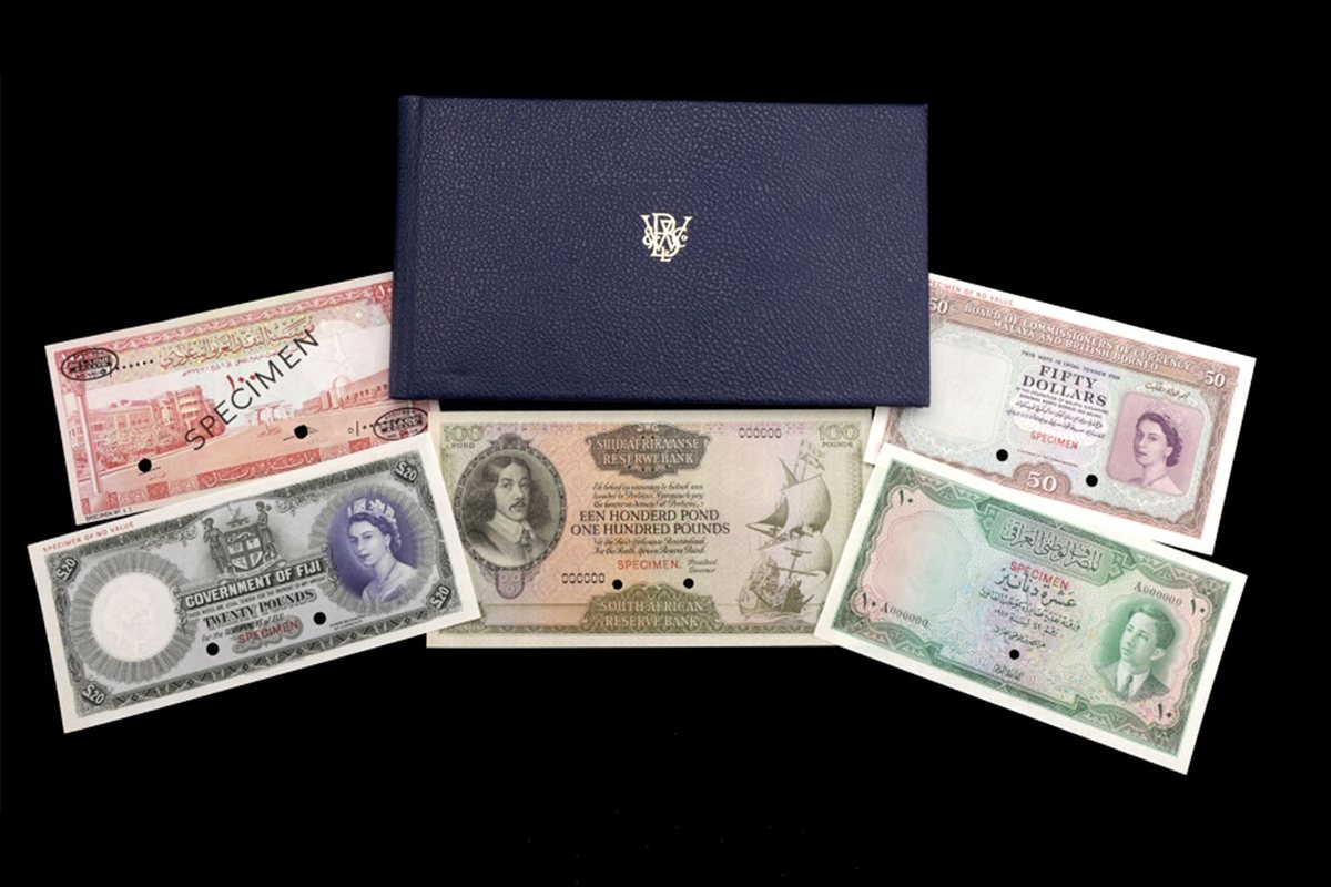 A dark blue leather album lies on a black background, surrounded by six banknotes stamped SPECIMEN with holes punched through them: notes from Fiji, Malaya and British Borneo, South Africa, Iraq and Saudi Arabia, two of them with portraits of a young Queen Elizabeth II.