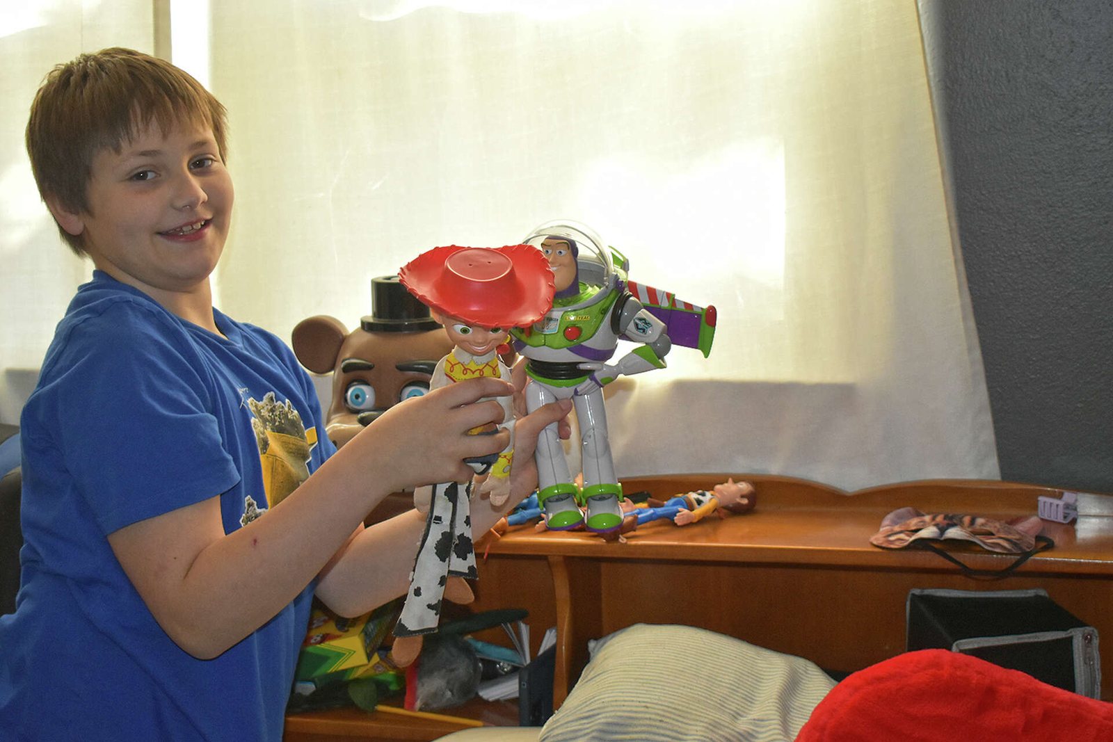 A smiling boy in a blue T-shirt holding Toy Story figures of Jessie and Buzz Lightyear.