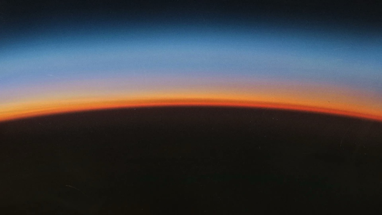 A curved horizon seen from high up, a thin band of orange and pale blue light along its edge and darkness above and below it