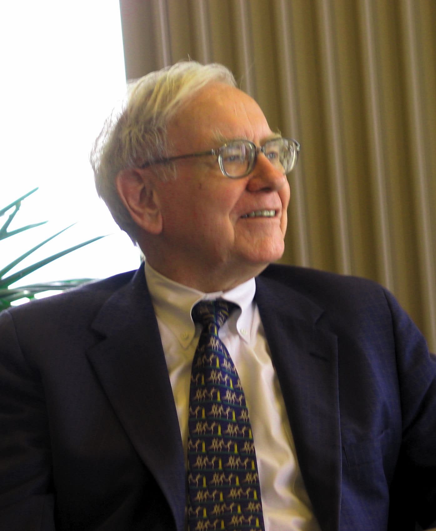 Warren Buffett in a dark suit and patterned tie, smiling and looking to one side, in front of a beige curtain