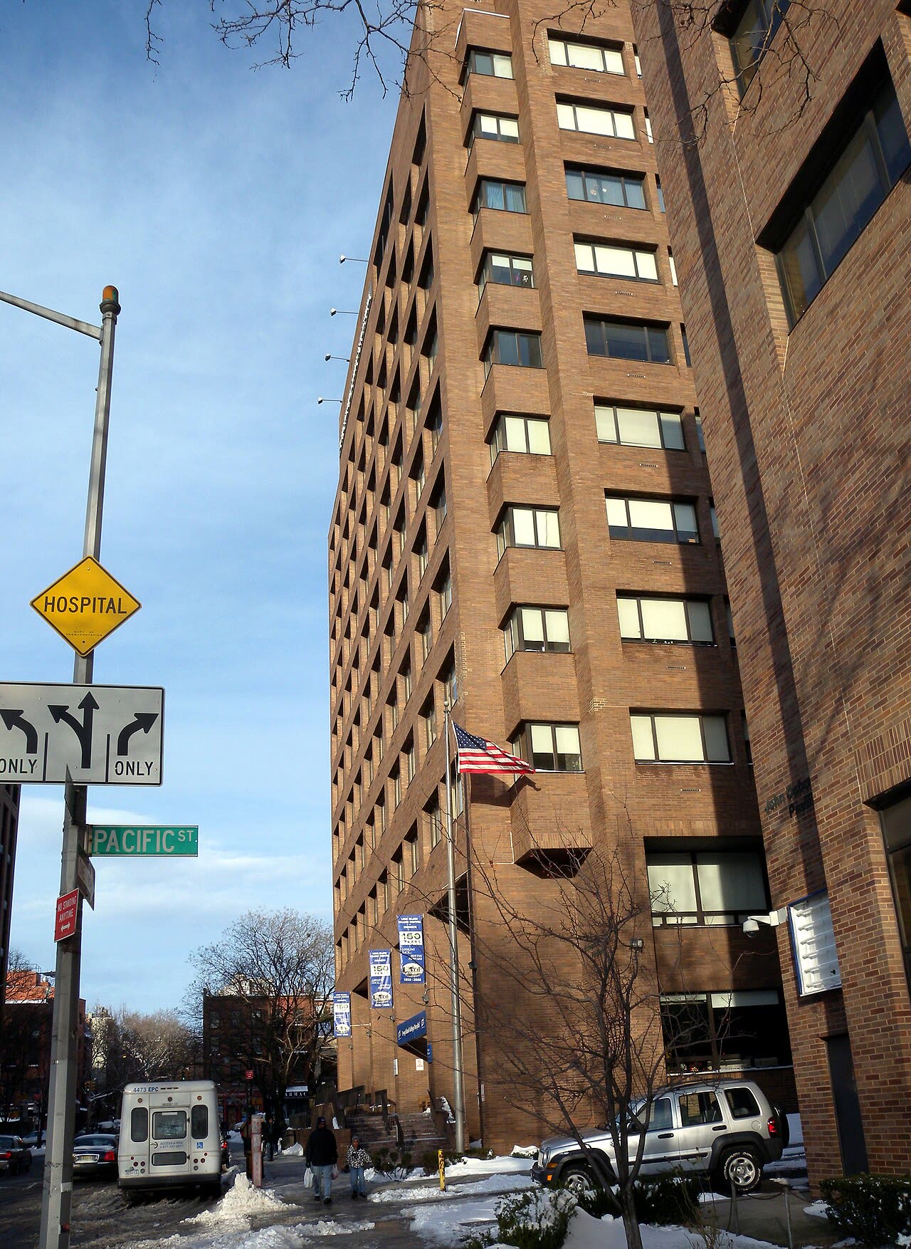A wide brick and glass hospital building on a corner in Brooklyn, photographed on a sunny afternoon with snow on the ground and bare trees along the street