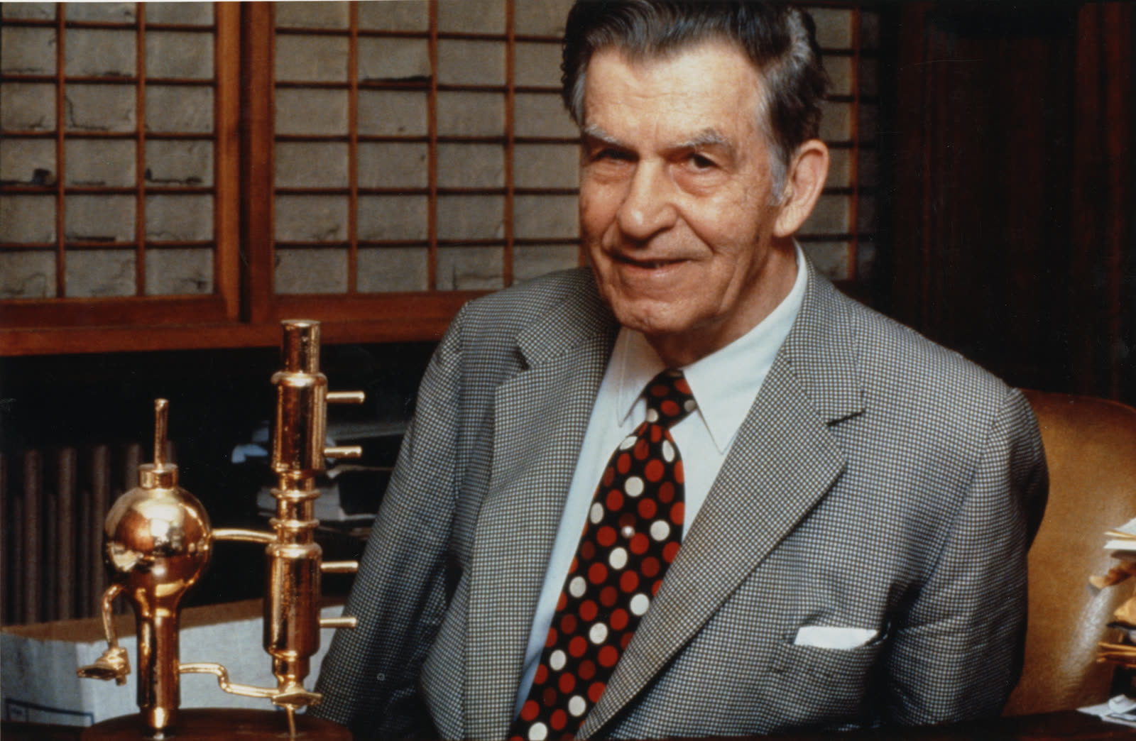 An elderly man in a checked jacket and red polka-dot tie smiles at the camera in a wood-panelled office, with a small gold-plated laboratory still on the desk beside him
