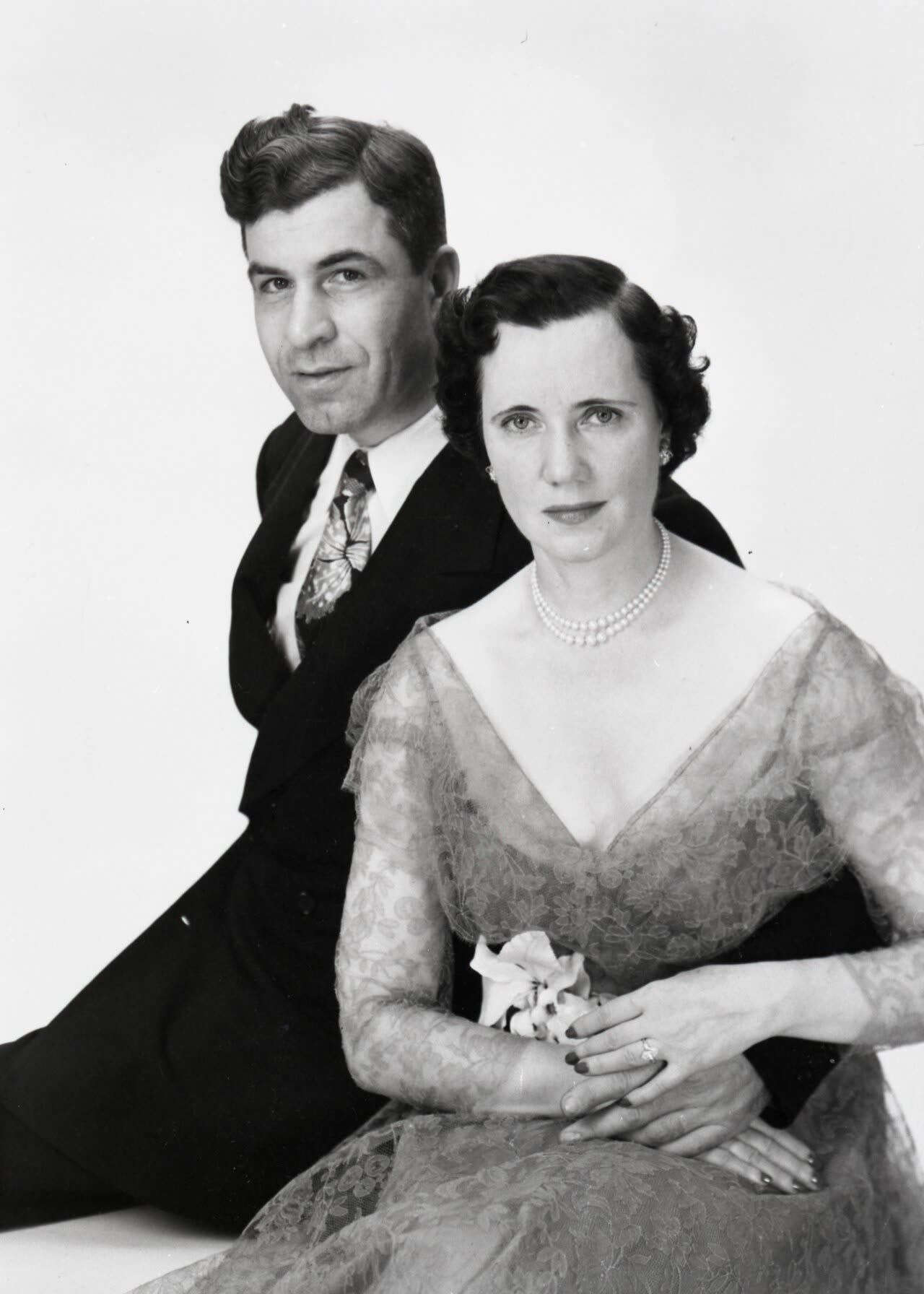 A black and white studio portrait of a man in a dark suit and patterned tie sitting behind a woman in a lace dress and pearls, his arm around her waist, both looking at the camera