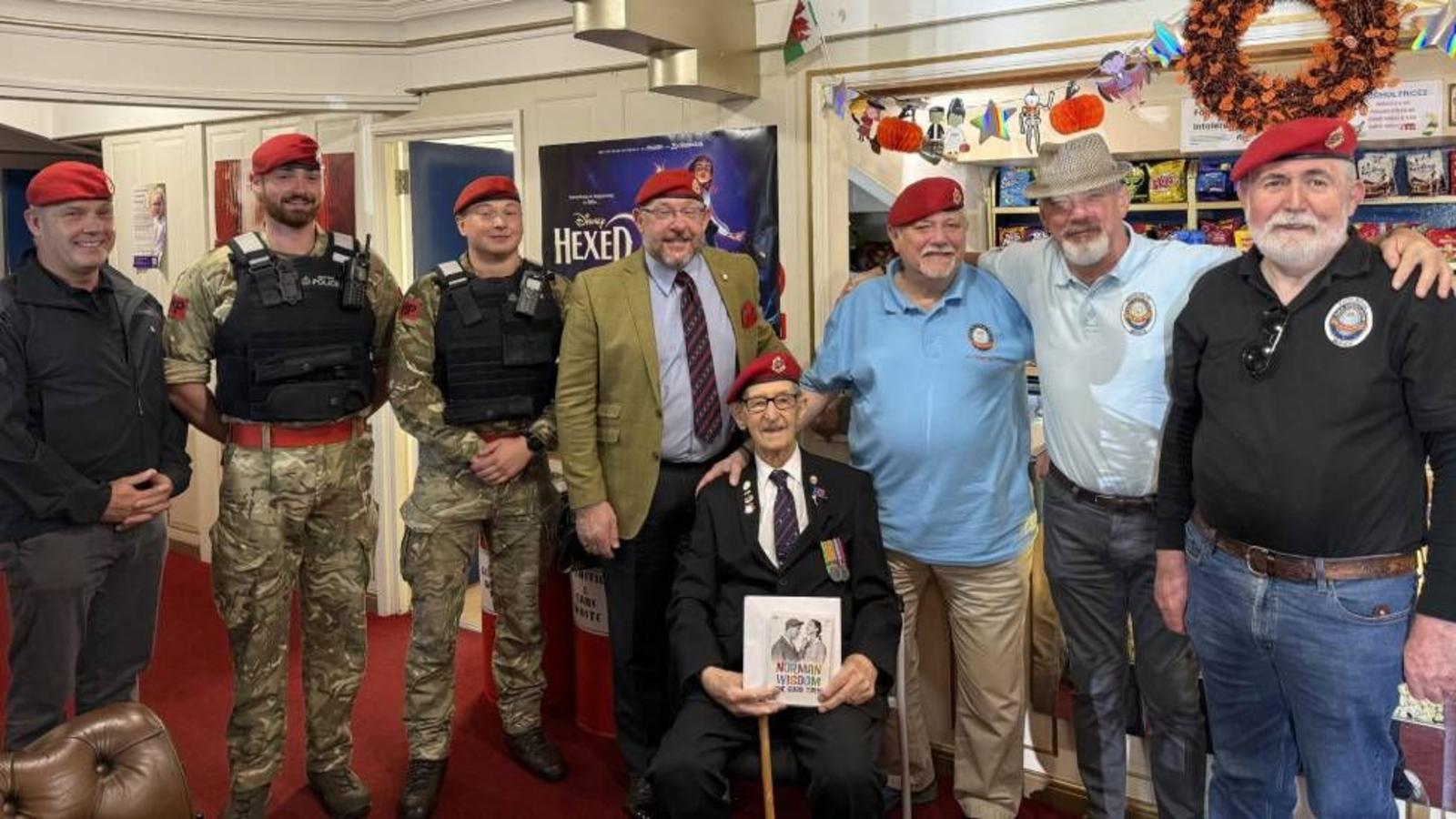Owen Filer sits in a dark suit and red beret, holding a framed photo, surrounded by soldiers and veterans in red berets in a room decorated with bunting.