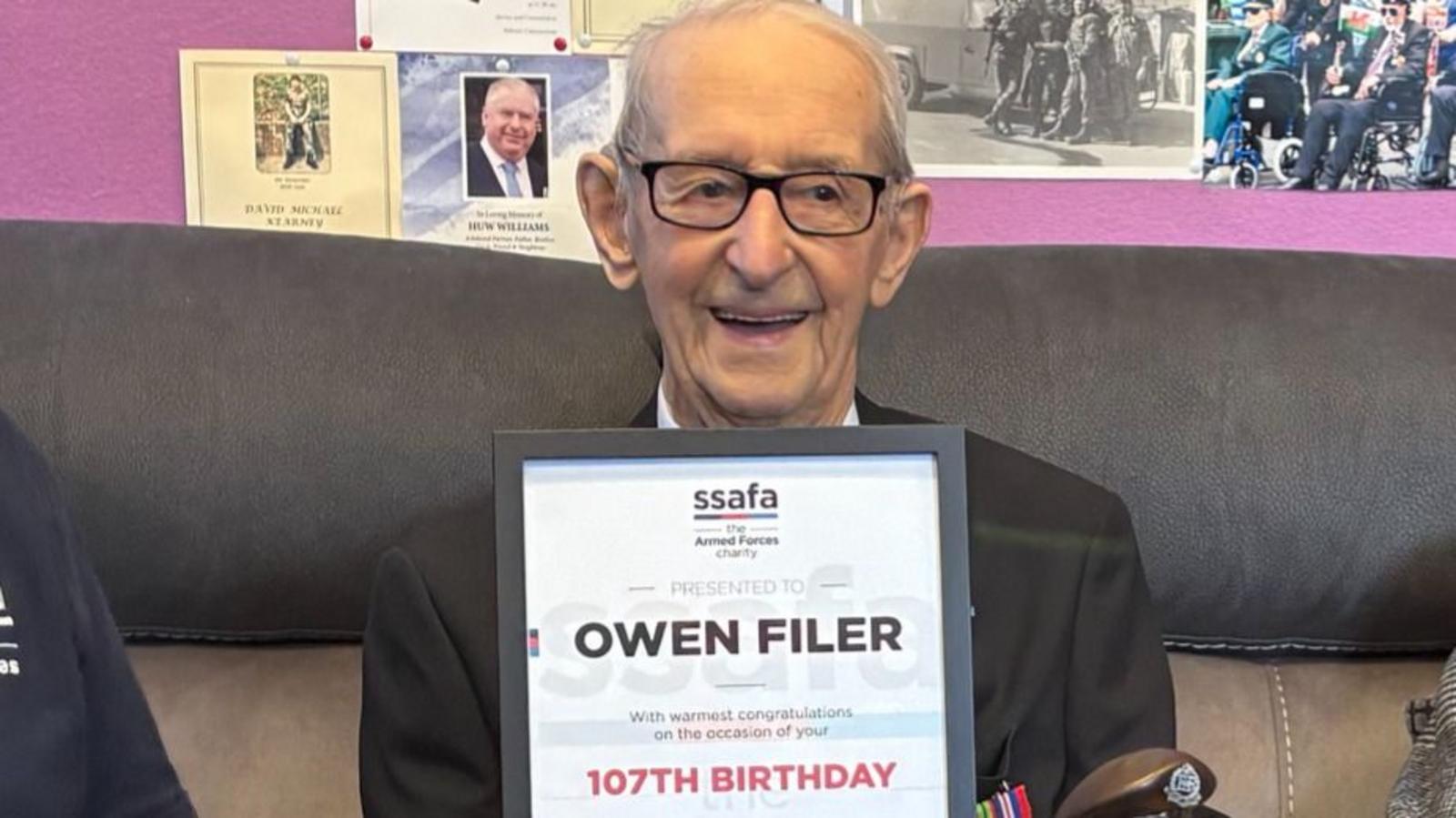 Owen Filer, a smiling 107-year-old man in glasses, a dark blazer and medals, holds a framed SSAFA certificate with his name and the words 107th birthday.