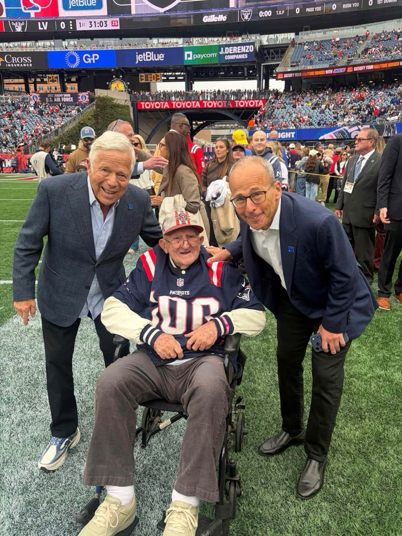 An old man in a Patriots shirt with the number 100 sits in a wheelchair on the field, with Robert Kraft on his right and Jonathan Kraft on his left.