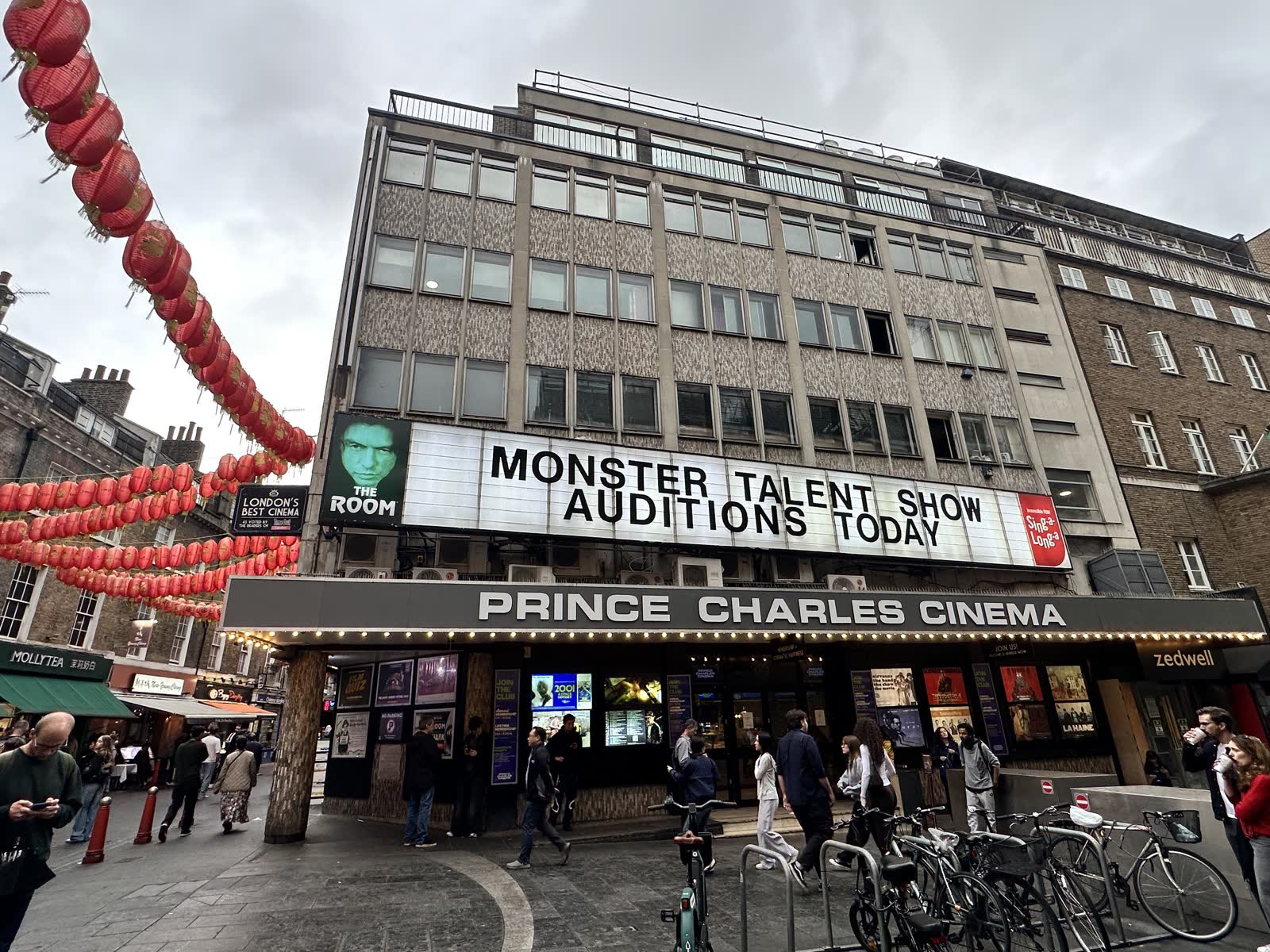 The front of the Prince Charles Cinema on a grey day, with a white letter board reading Monster Talent Show Auditions Today, a poster for The Room, red lanterns on the left and a Zedwell sign on the right.