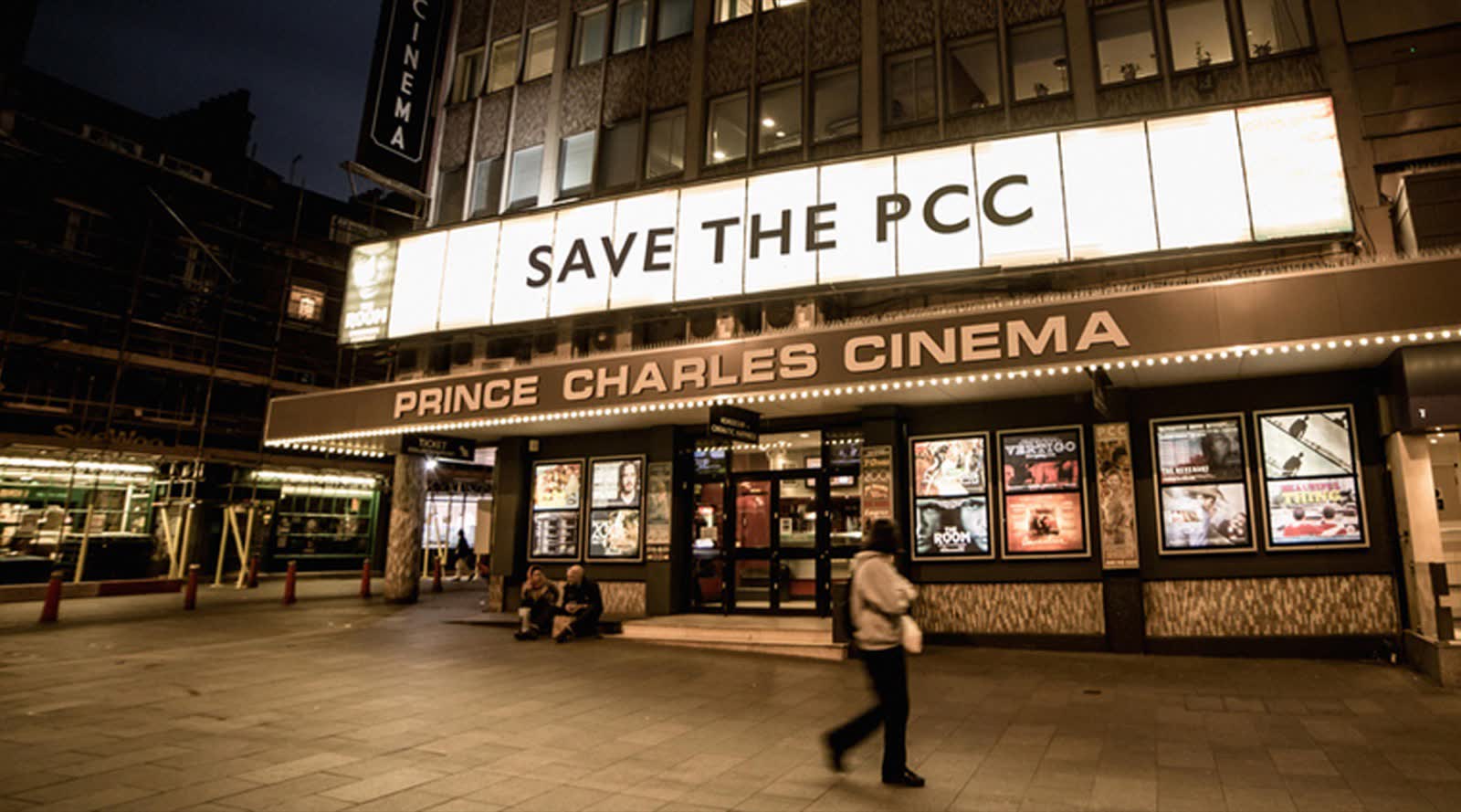The cinema at night with its letter board reading Save the PCC, posters for The Room and Vertigo in the windows, and two people sitting on the step.