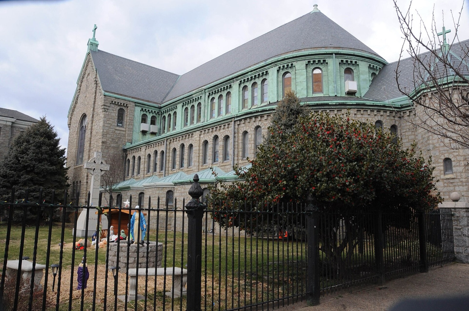 A large grey stone church with green copper trim and a curved apse, seen over an iron fence with a nativity scene and a stone cross in the garden